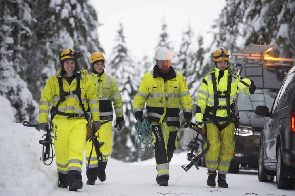 El-kraft Four OneCo workers in walking through the snow in their uniforms with various equipment. Two vehicles can be seen in the background.