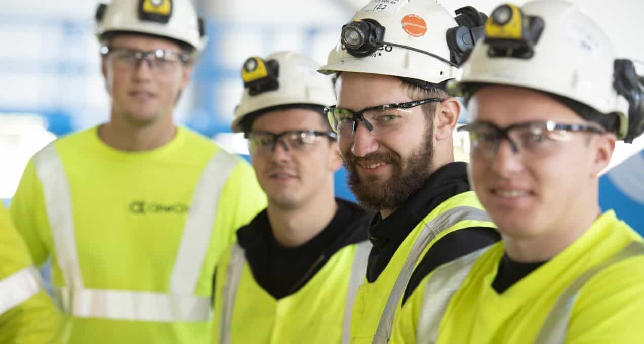 Fire OneCo-ansatte smiler mot kameraet, iført arbeidstøy, hjelmer og briller.