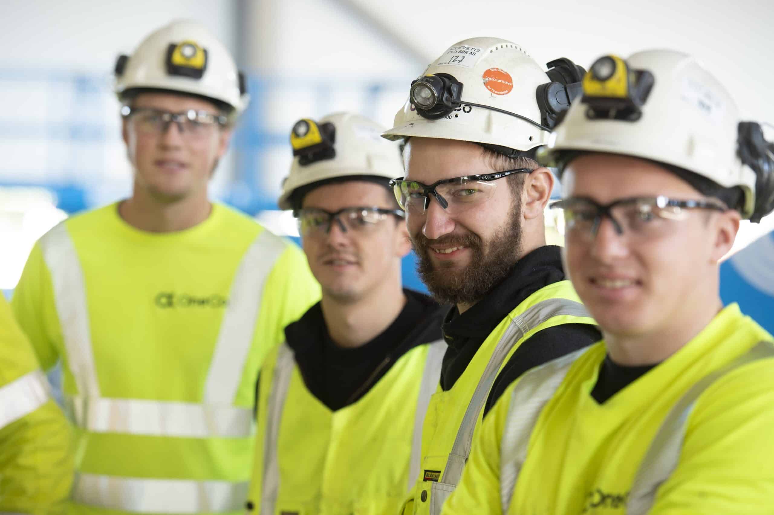 Fire OneCo-ansatte smiler mot kameraet, iført arbeidstøy, hjelmer og briller.