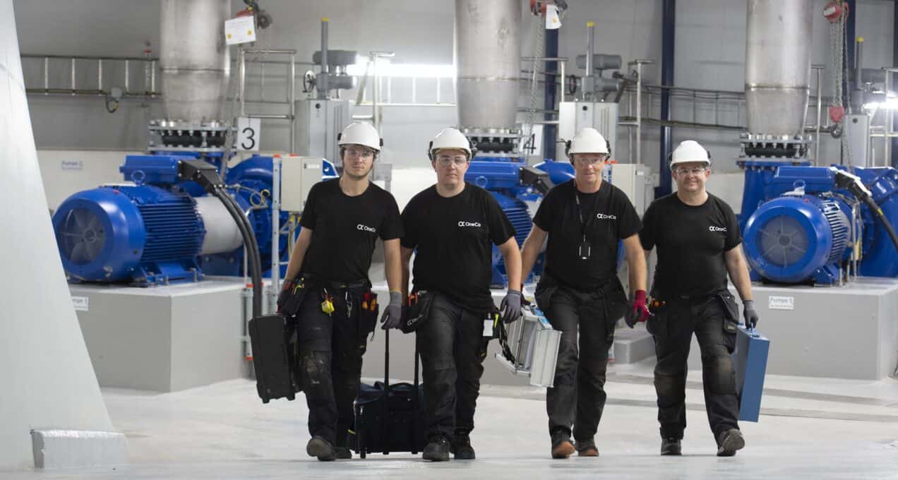 A team of four people is walking in a line towards the camera whilst carrying their electric equipment. A professional production system is visible in the background.