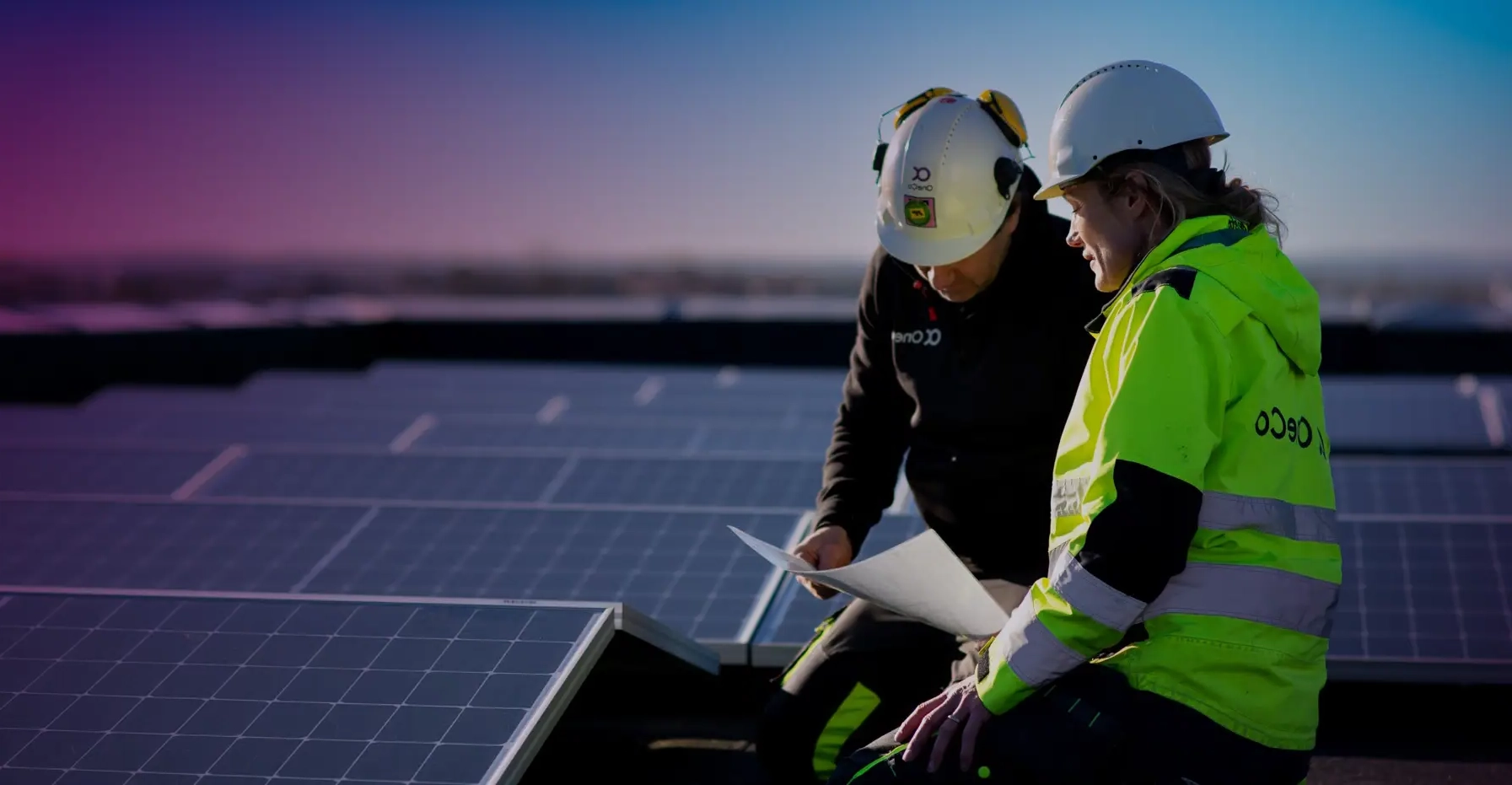 Two OneCo workers look together at a paper whilst seated next to a large solar system installation