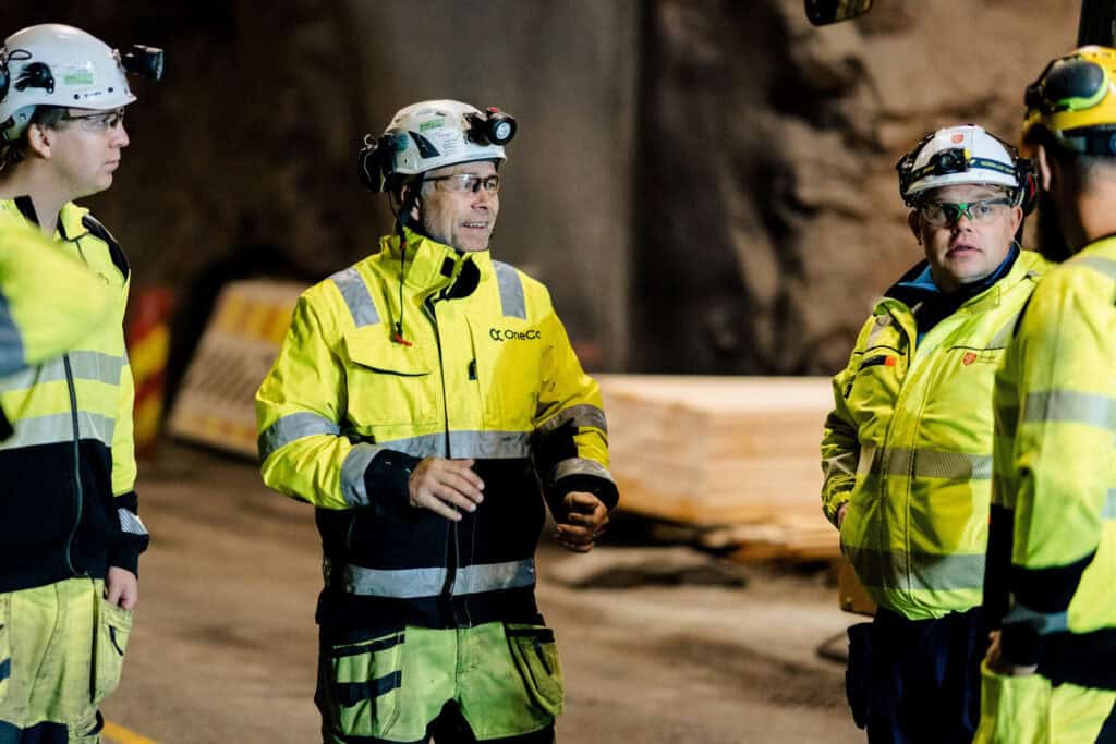 A group of OneCo workers are seen meeting together on the job with helmets and uniforms on.
