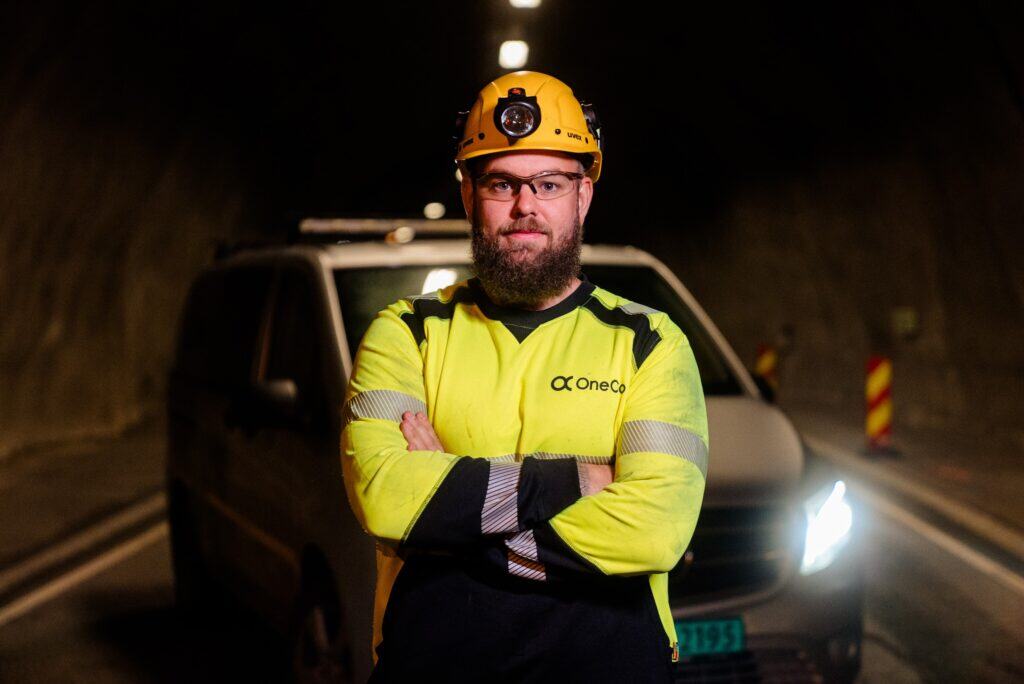 A OneCo worker is posing in front of the camera with work clothes on. They are in a tunnel with their car parked directly behind them.