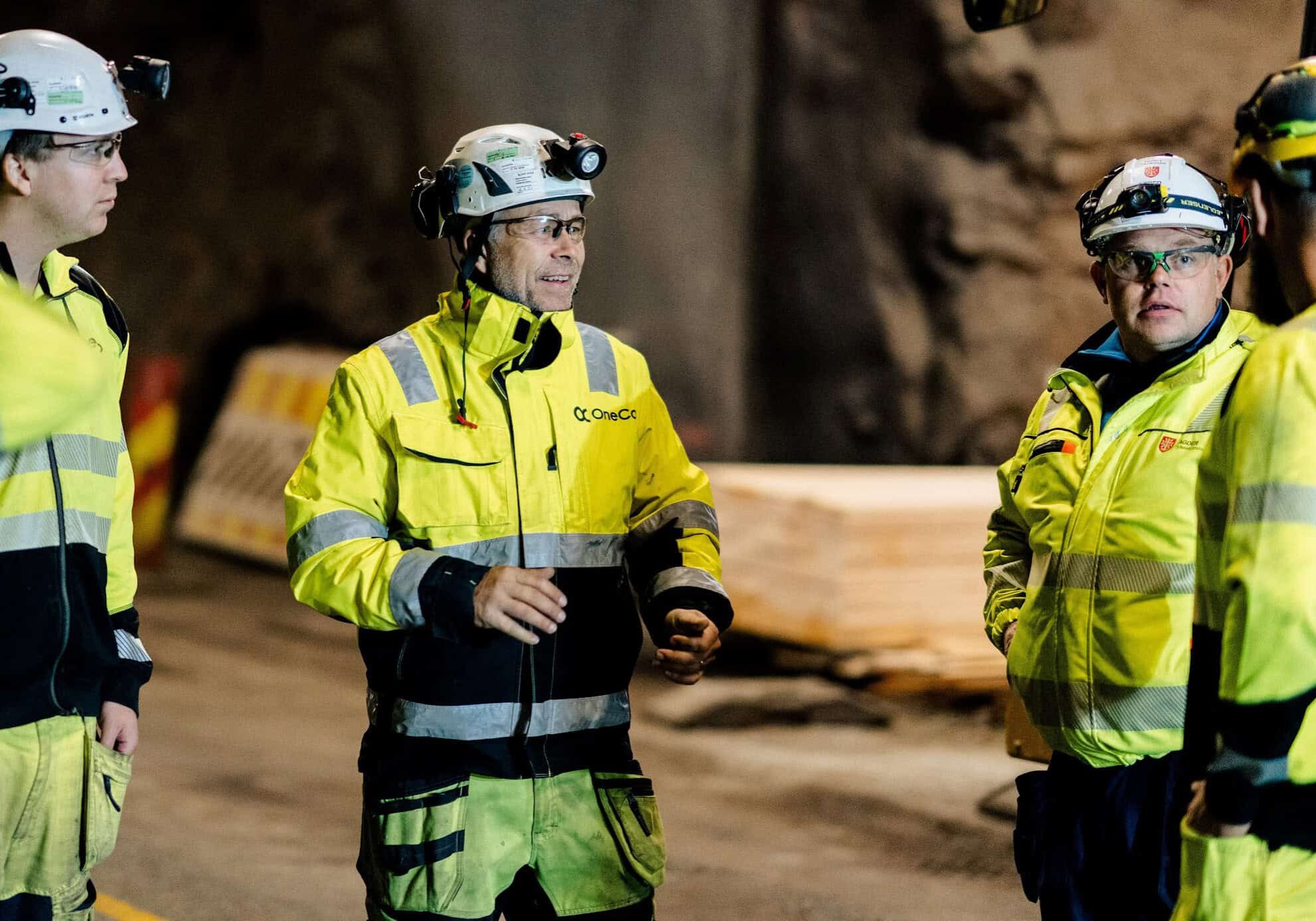 A group of OneCo workers are seen meeting together on the job with helmets and uniforms on.