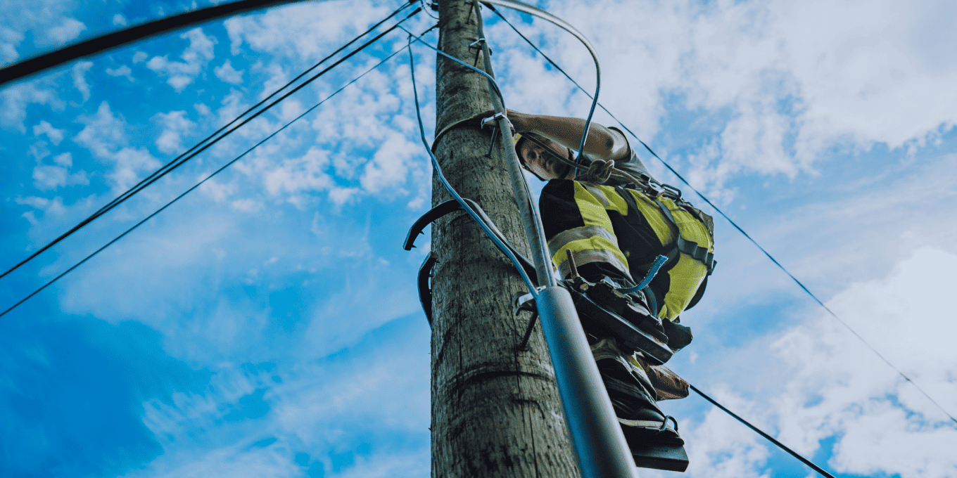 OneCo worker working up on a telephone pole, photo taken from below with blue sky and clouds visible in the background